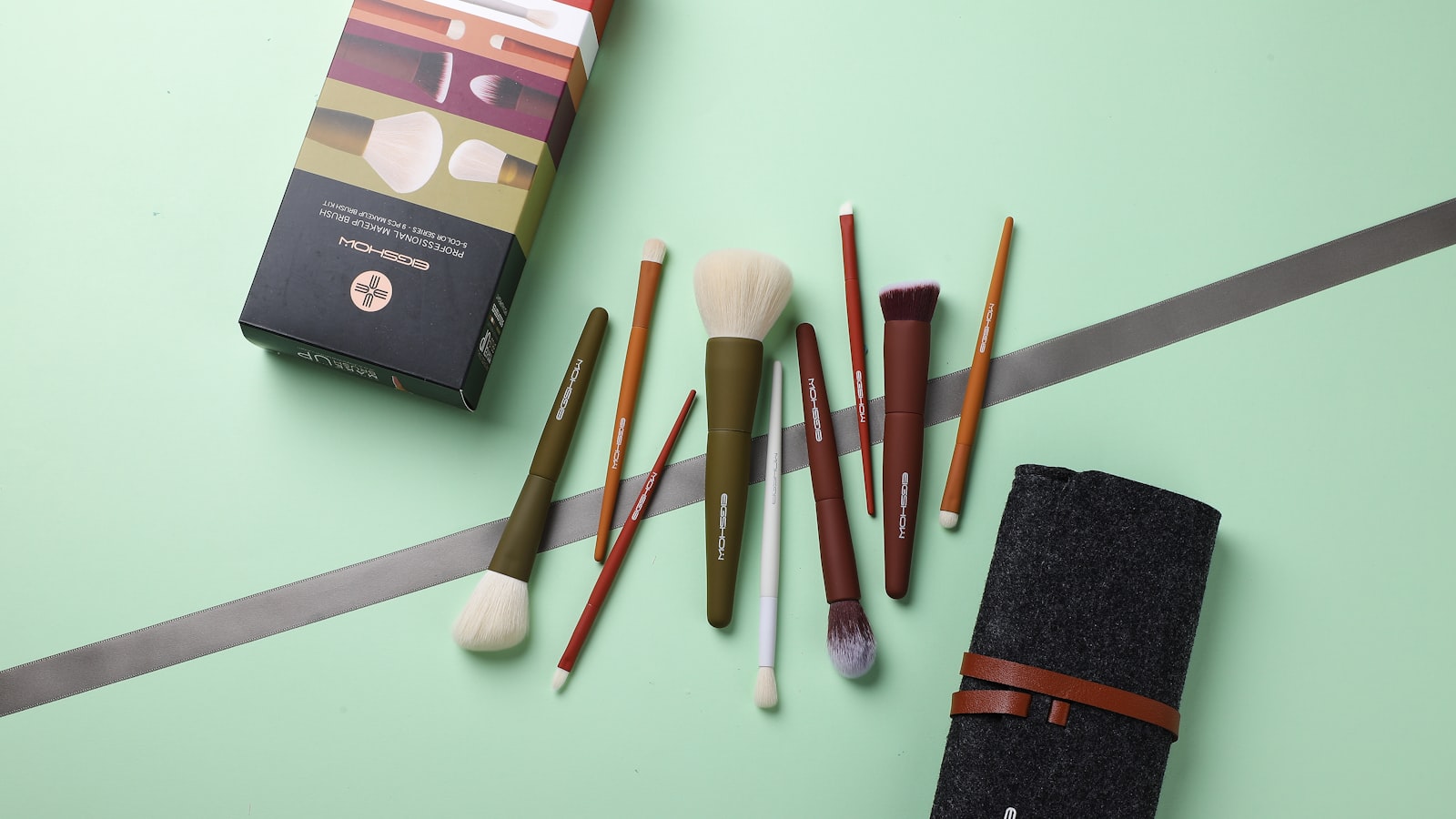 Nine makeup brushes are arranged on a green surface alongside a box, a grey ribbon, and a felt case