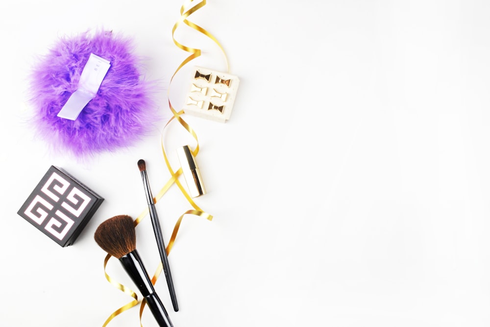 Makeup brushes, a purple puff, a patterned box, and earrings sit on a white surface with gold ribbon