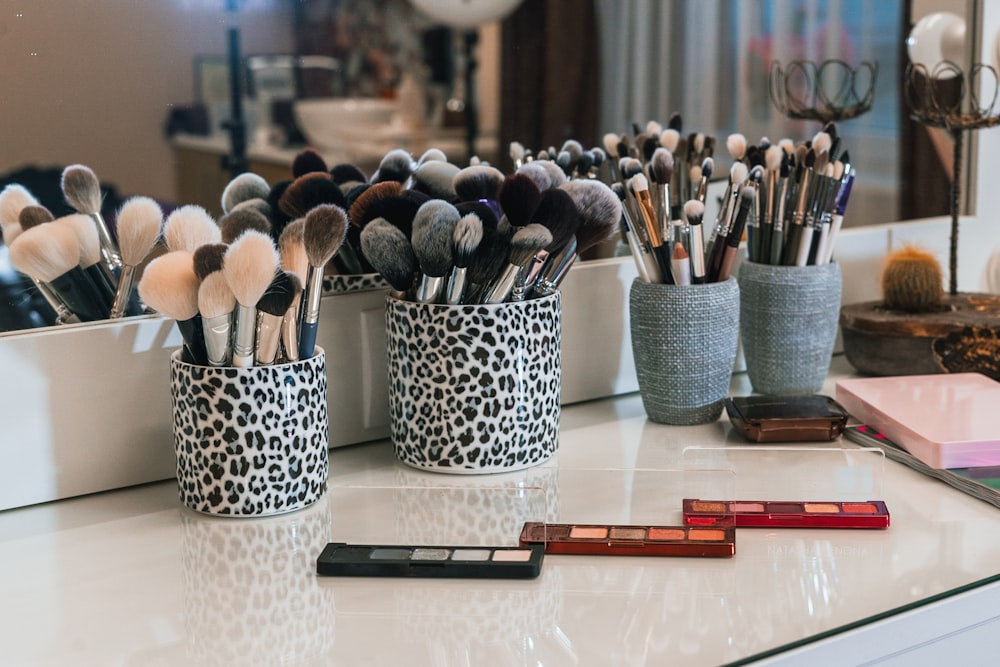 Containers of makeup brushes and palettes are arranged on a white dressing table with a mirror