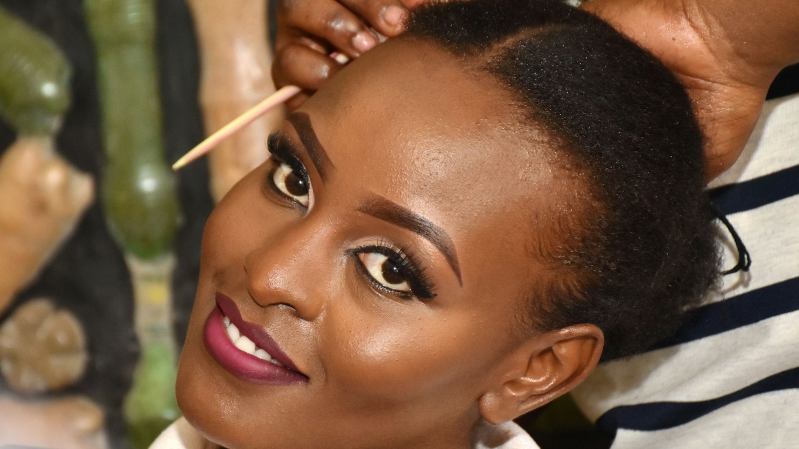 Close-up of a Black woman with dark hair and lipstick looking up while someone styles her hair