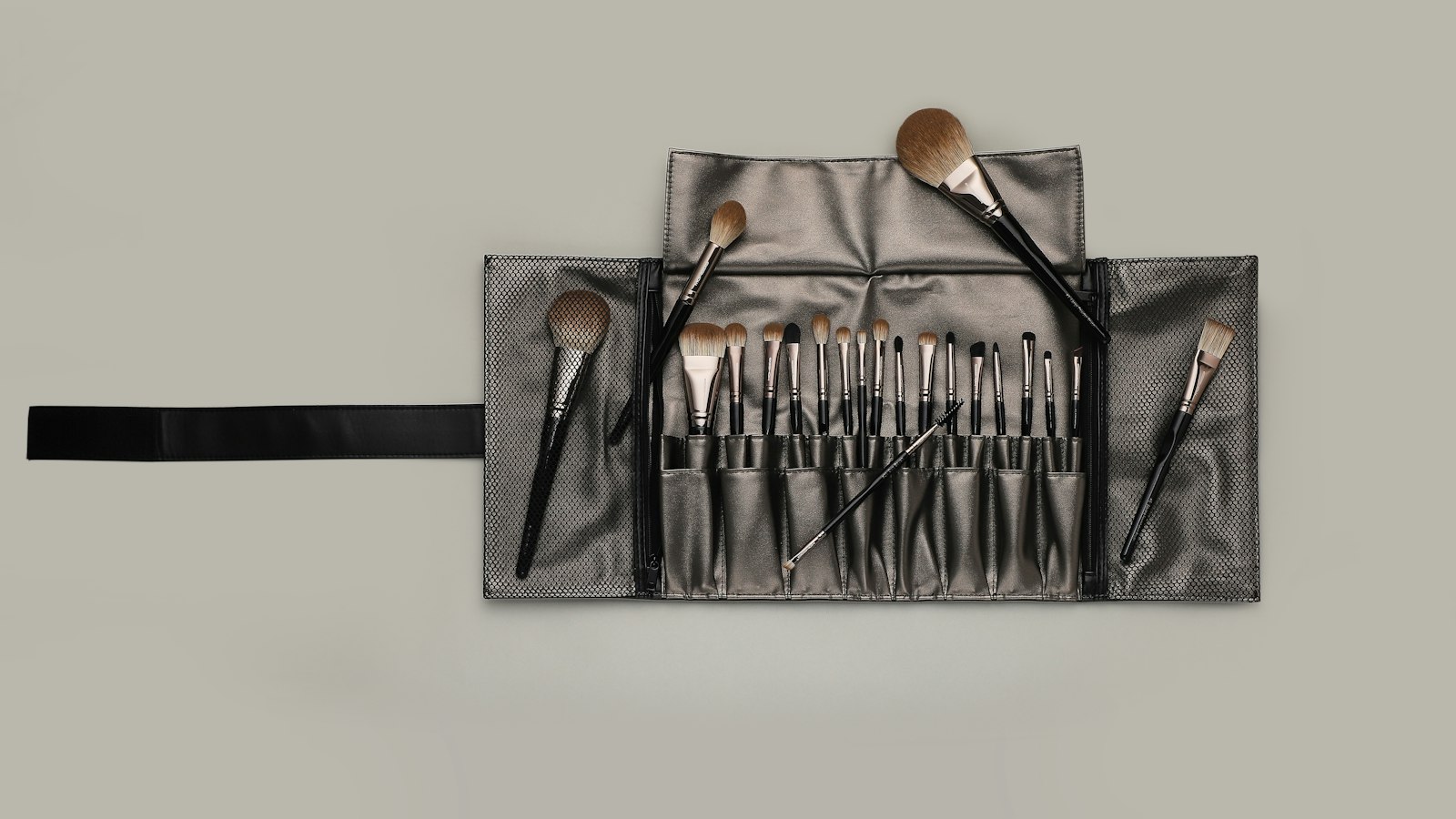 A roll-up makeup brush case contains a set of brushes with black handles and brown bristles