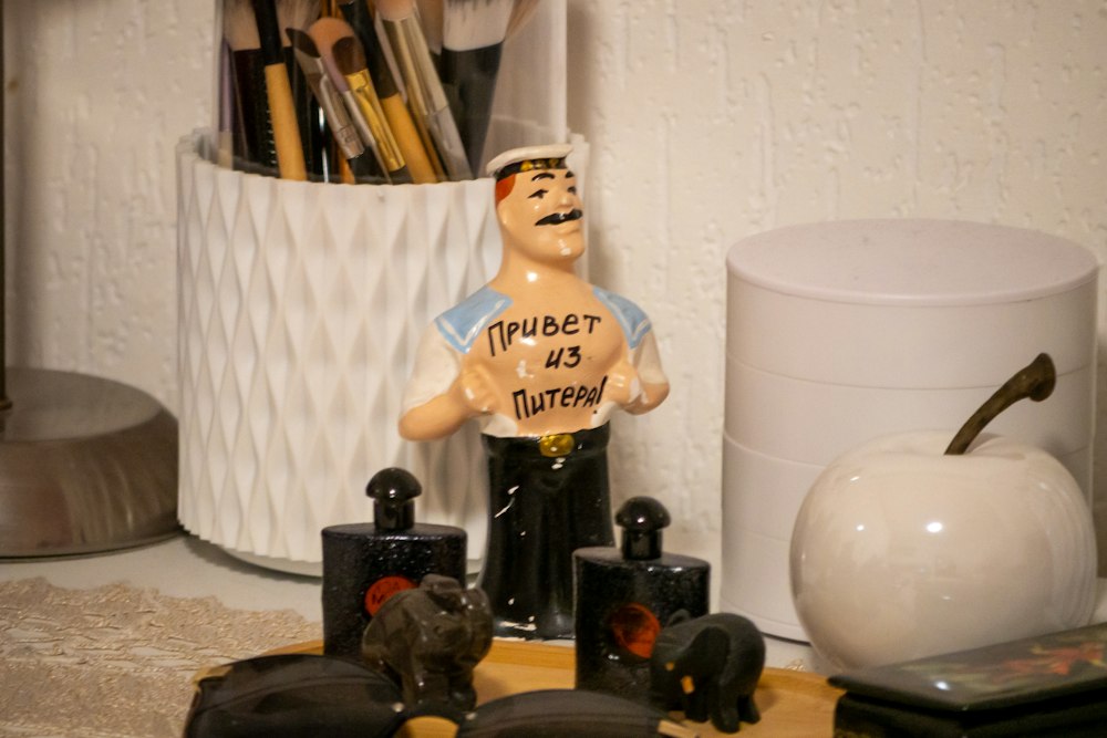 A small ceramic figure of a sailor stands surrounded by vanity items on a white surface