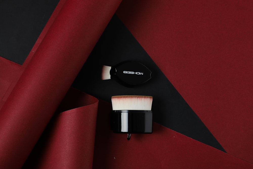 Two black makeup brushes are arranged on a dark background framed by red paper rolls