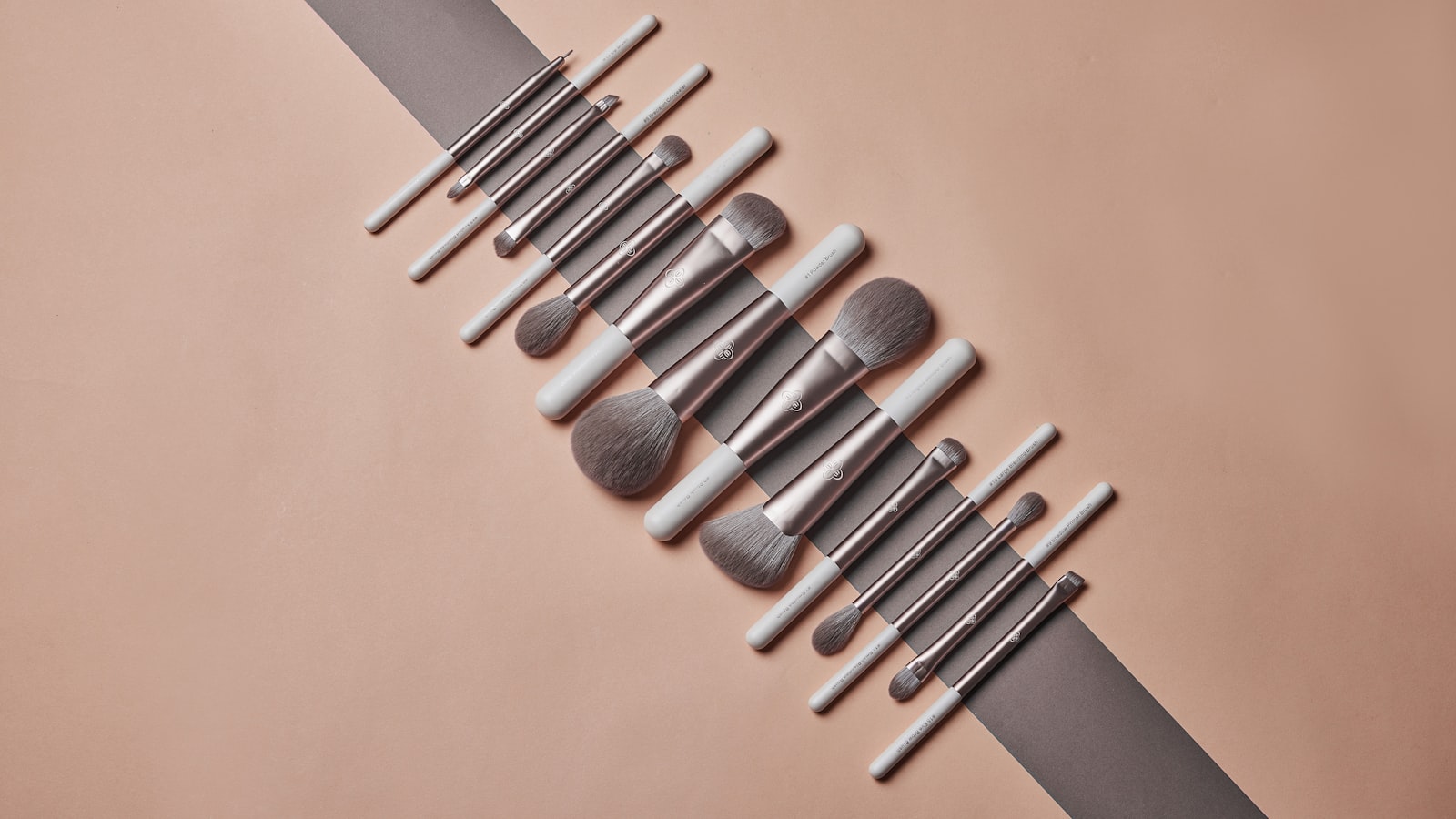 A collection of twelve makeup brushes are lined up diagonally on a gray ribbon against a peach background