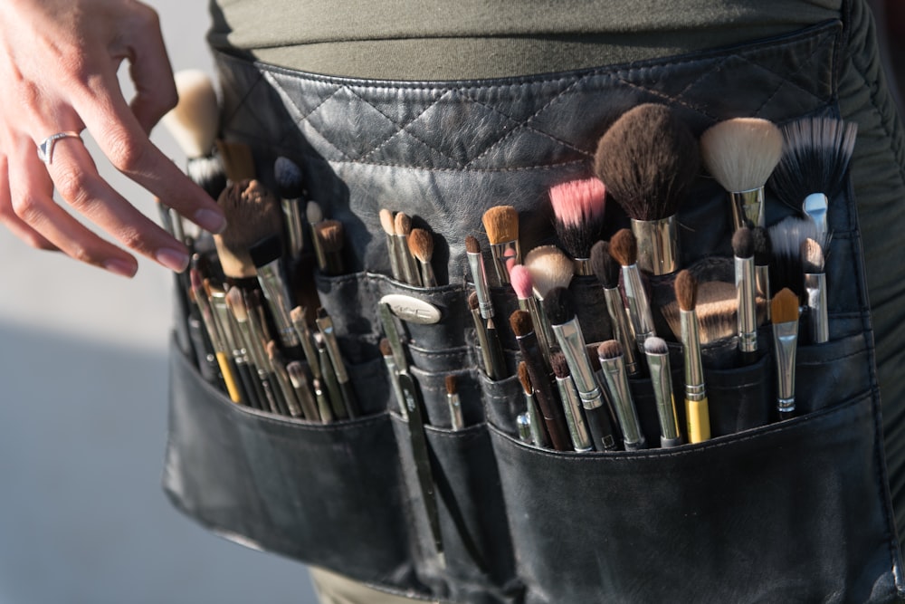 A person wearing a black utility belt filled with various makeup brushes