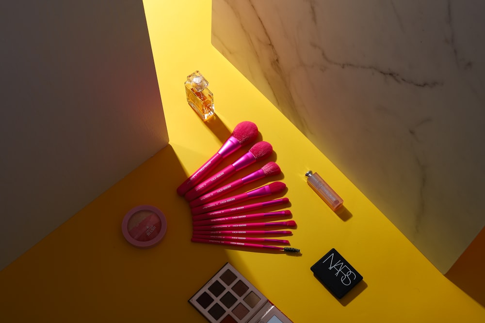 Cosmetics, including pink brushes, a palette, and a perfume bottle, are arranged on a yellow surface