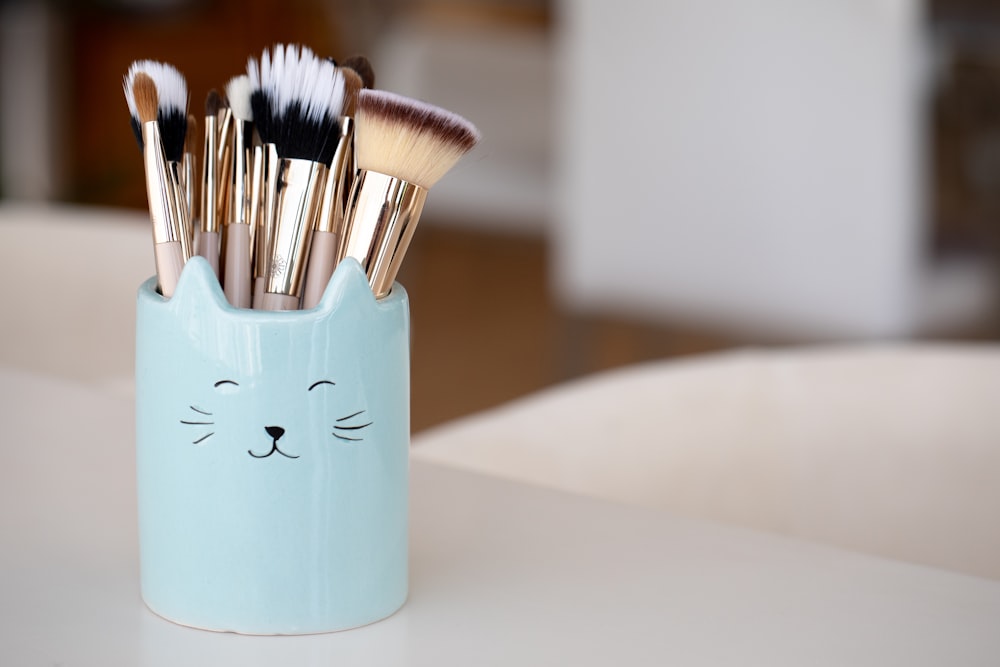 A light blue cat-shaped cup filled with various makeup brushes sits on a white surface