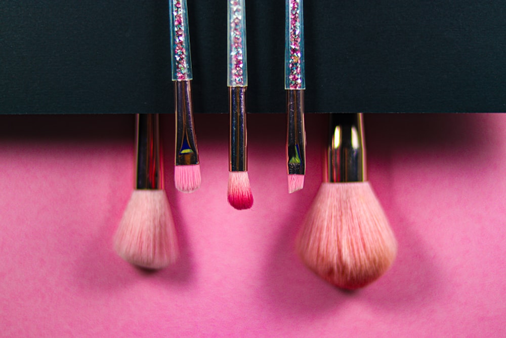 Five makeup brushes with pink bristles and glitter handles descend against a black and pink background