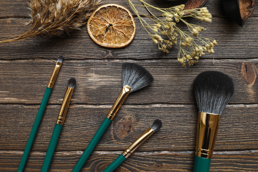 Five makeup brushes with green handles arranged on a wooden surface with dried plants and a dried citrus slice