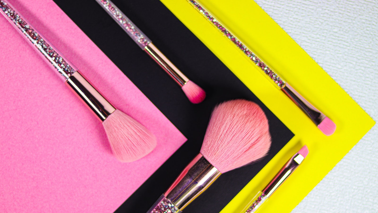 Four pink and silver makeup brushes are arranged on pink, black, and yellow paper