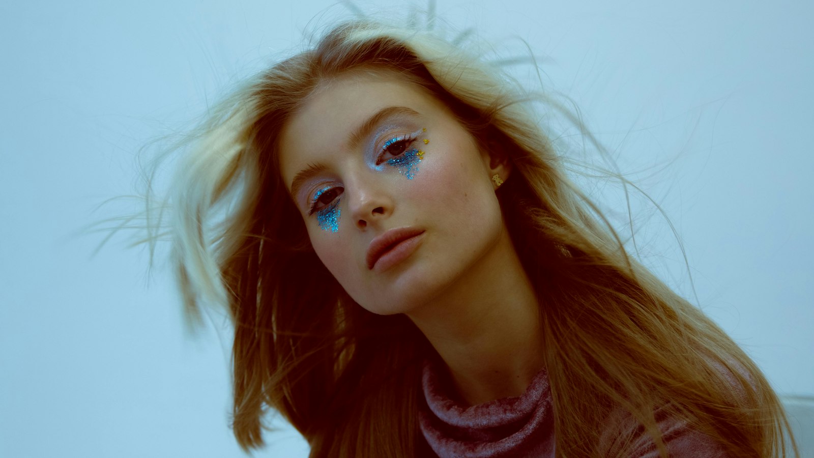 A close-up portrait of a woman with long blonde hair, blue glitter under her eyes, and a neutral expression