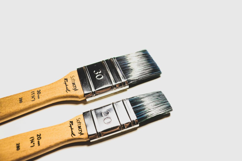 Two flat paint brushes with wooden handles lie diagonally on a white surface