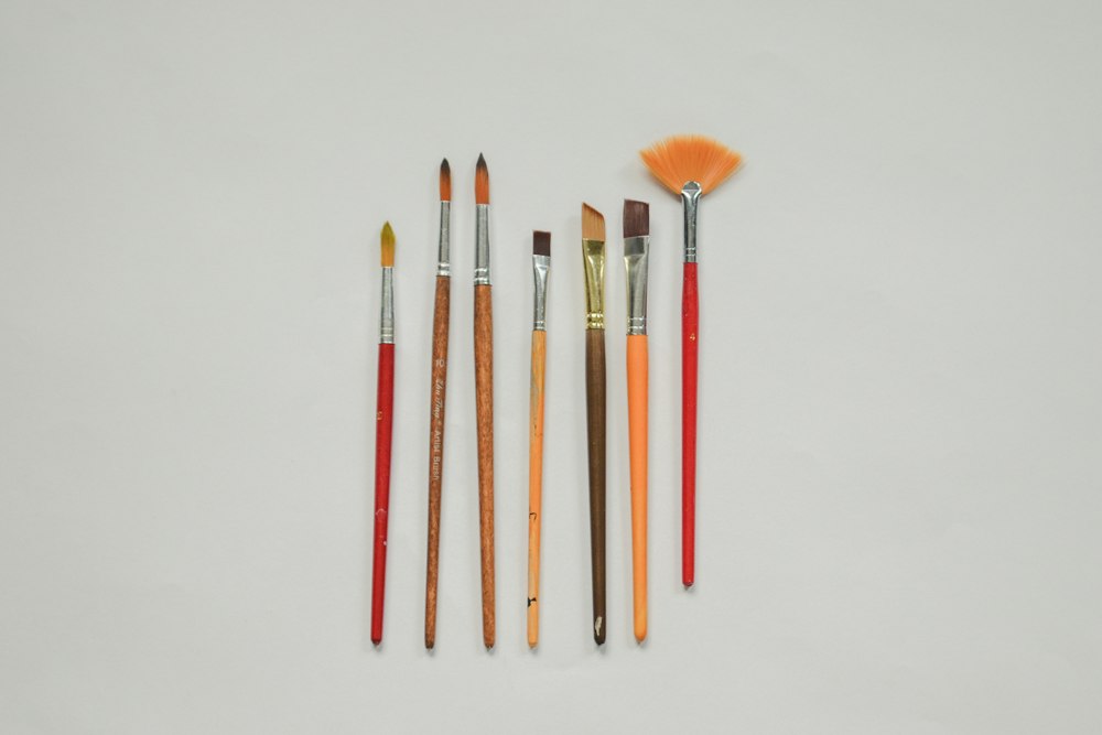 Seven assorted paint brushes lined up vertically on a plain surface