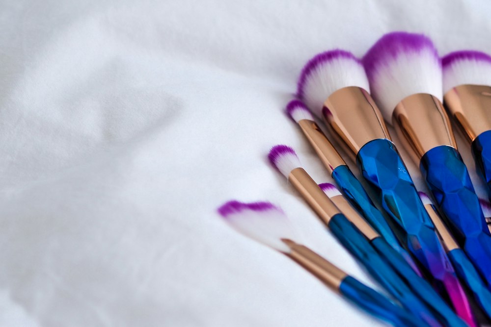 Seven makeup brushes with blue faceted handles and purple-tipped white bristles rest on white fabric