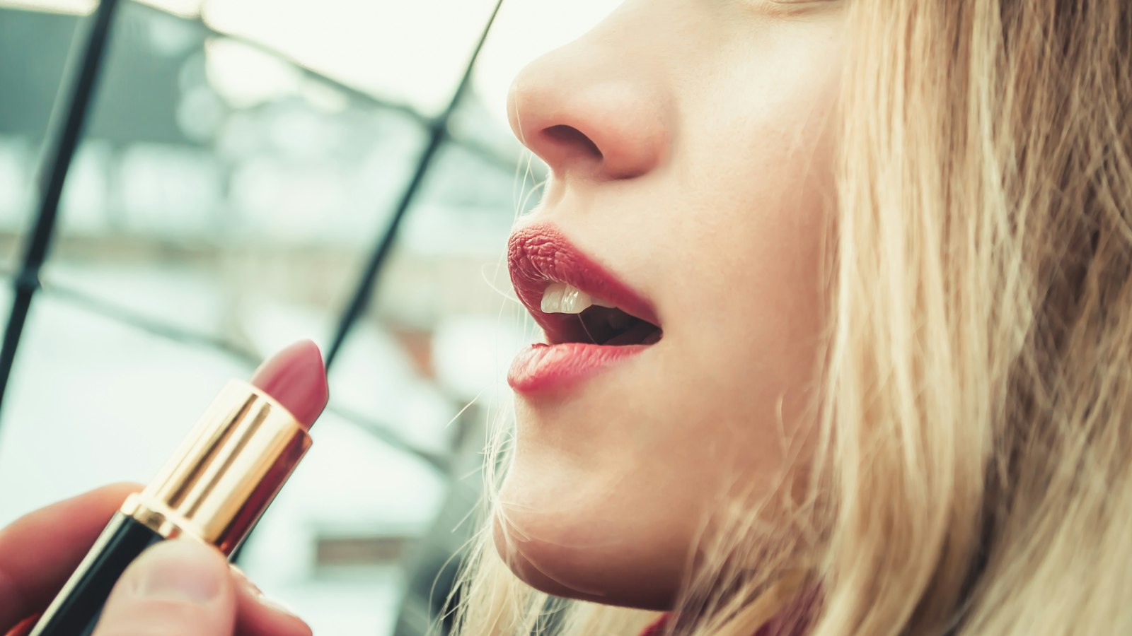 Close-up of a blonde woman holding a tube of lipstick near her open mouth