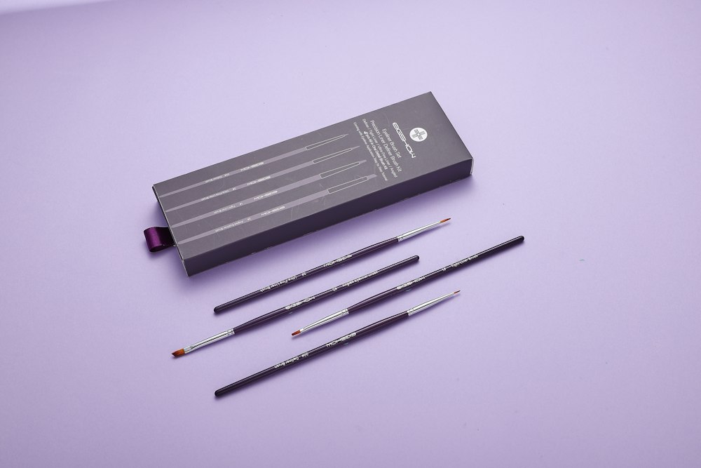 Four cosmetic brushes and their gray packaging box on a light purple surface