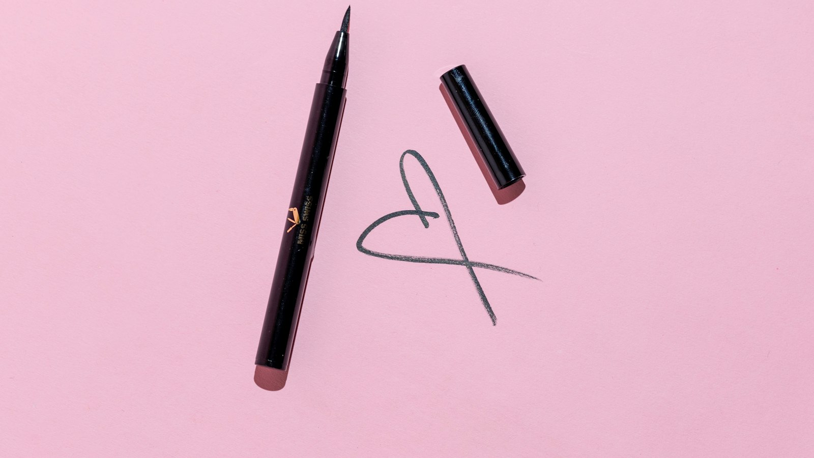 An open black eyeliner pen with its cap nearby lies on a pink surface next to a drawn heart