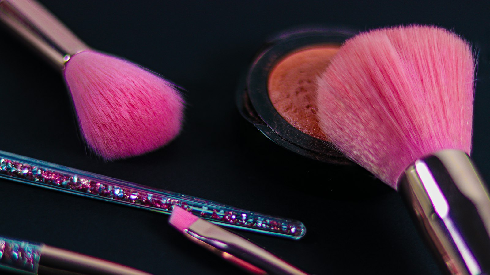 Close-up of makeup brushes with bright pink bristles, a compact with brown powder, and a glitter-filled handle