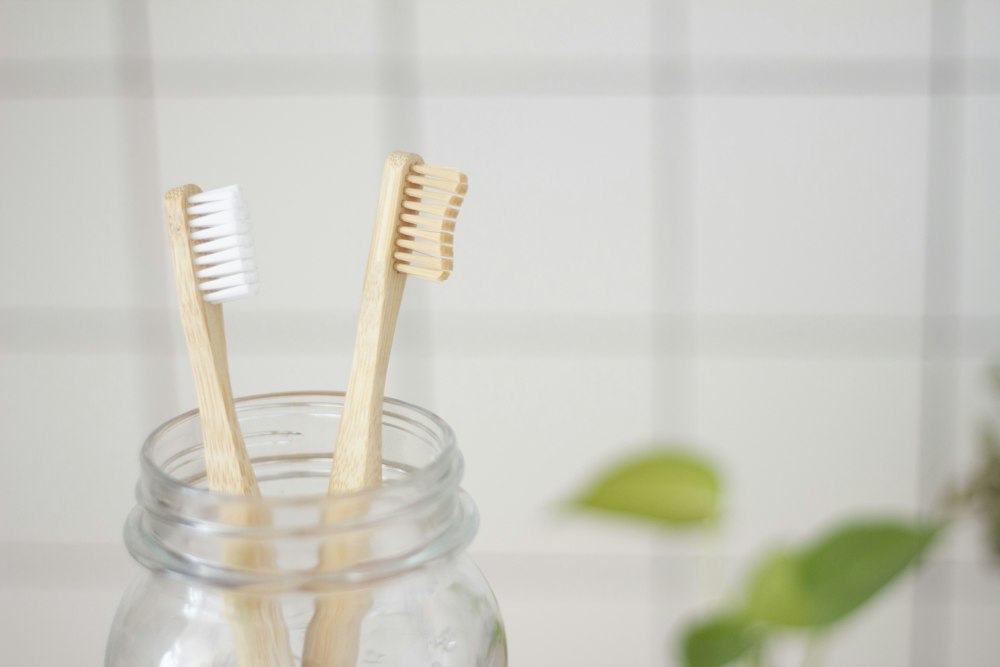 Two wooden toothbrushes in a glass jar against a white tile wall