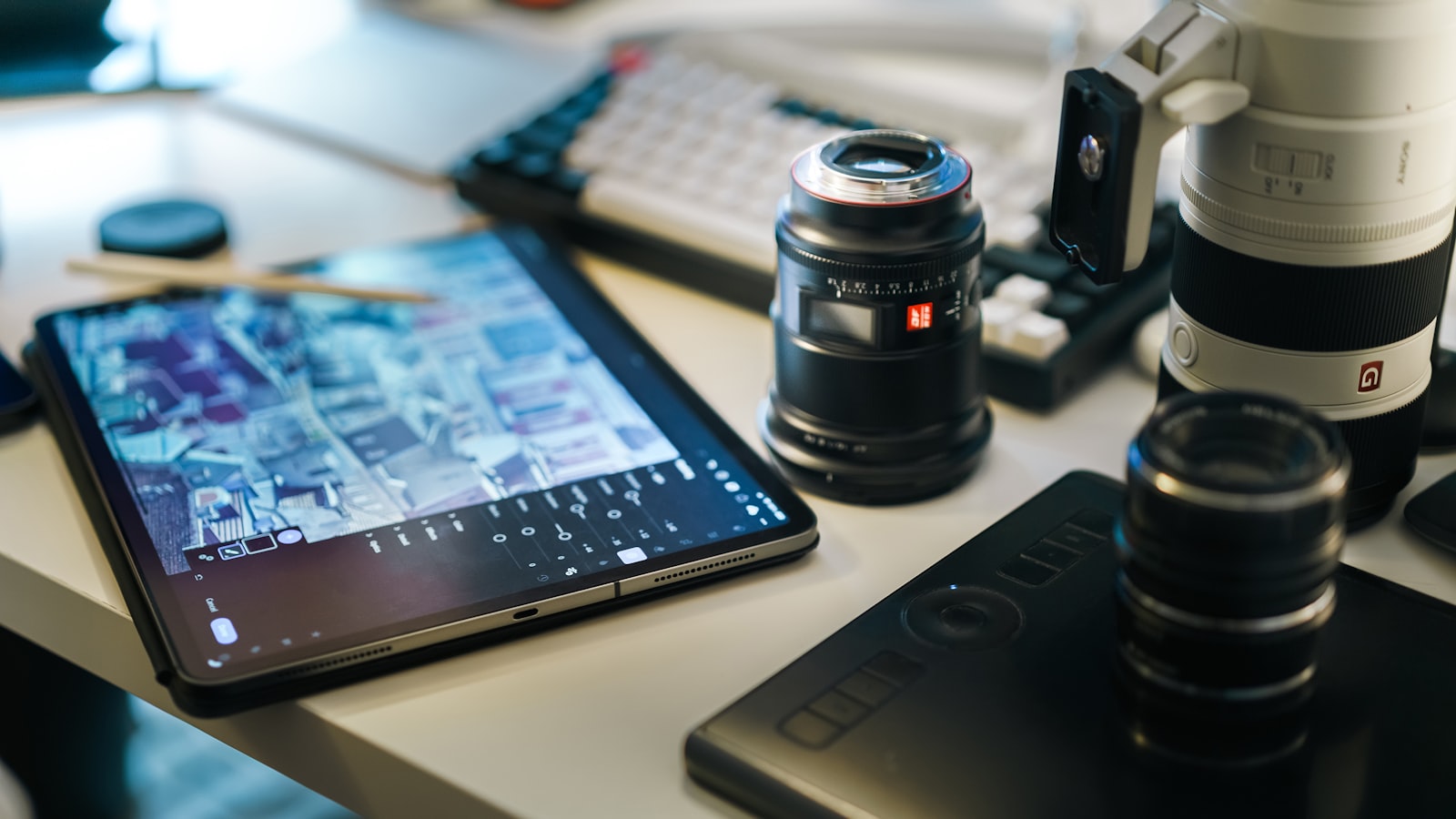 A tablet, camera lenses, and a keyboard are arranged on a desk