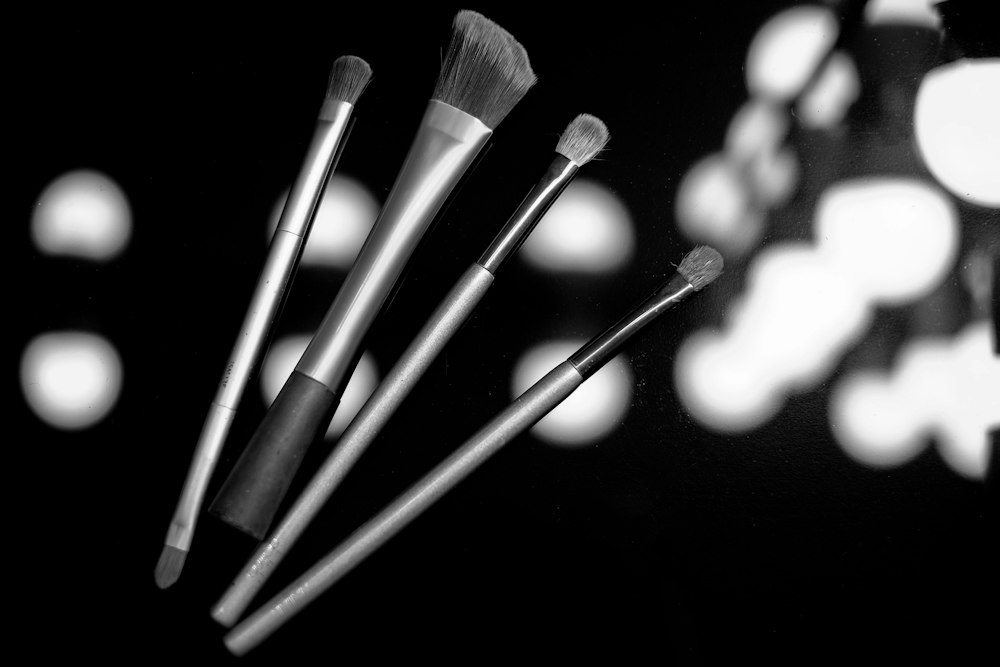 Four makeup brushes lie diagonally on a dark surface in this black-and-white photo