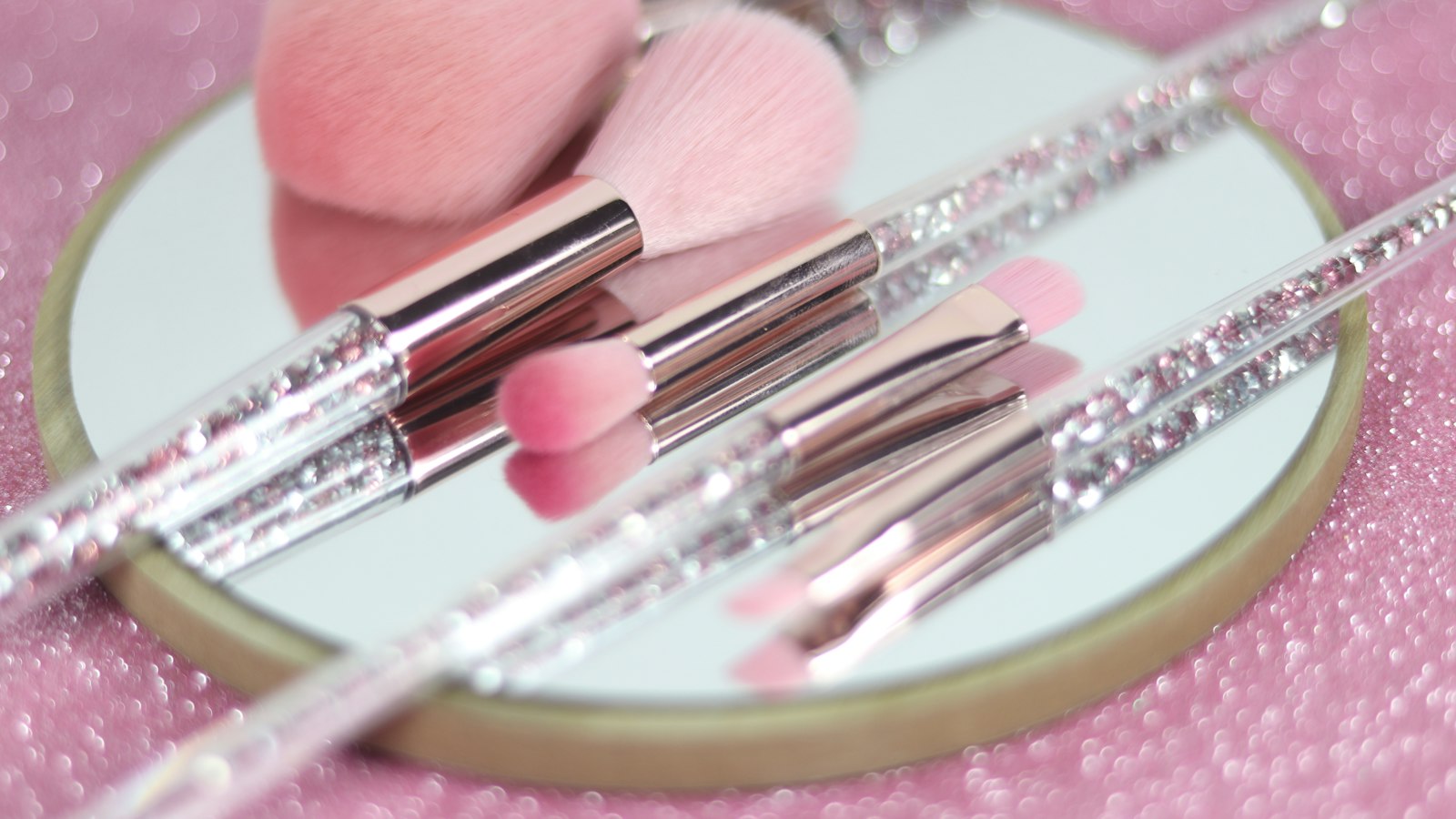 Pink cosmetic brushes with crystal-filled handles lie on a round mirror against a pink glittery background