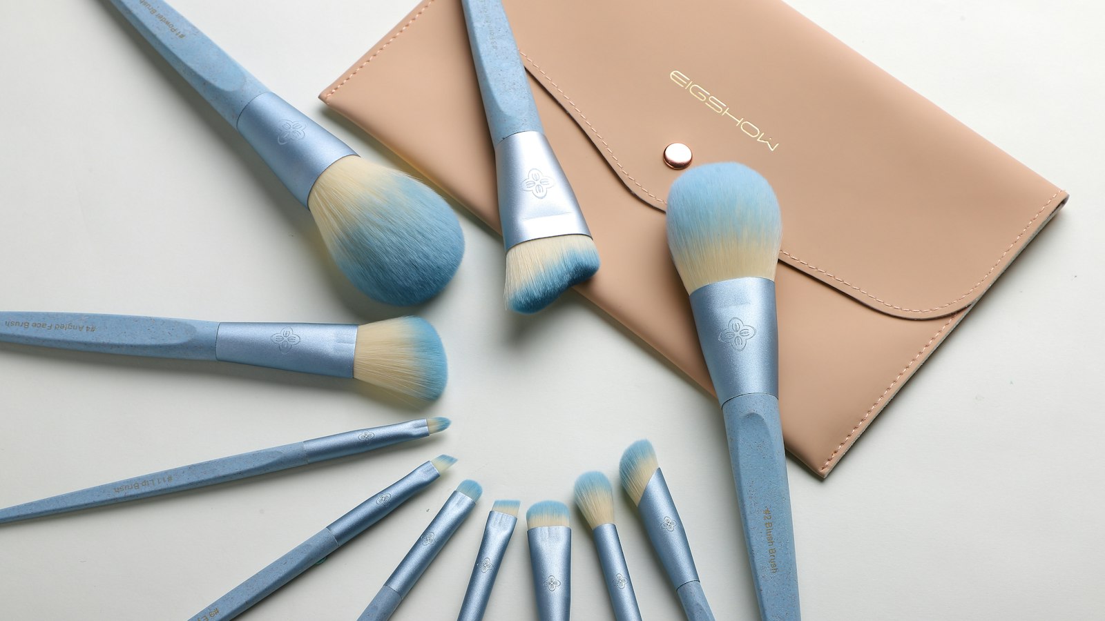 Light blue makeup brushes are arranged in a circle on a white surface next to a tan case